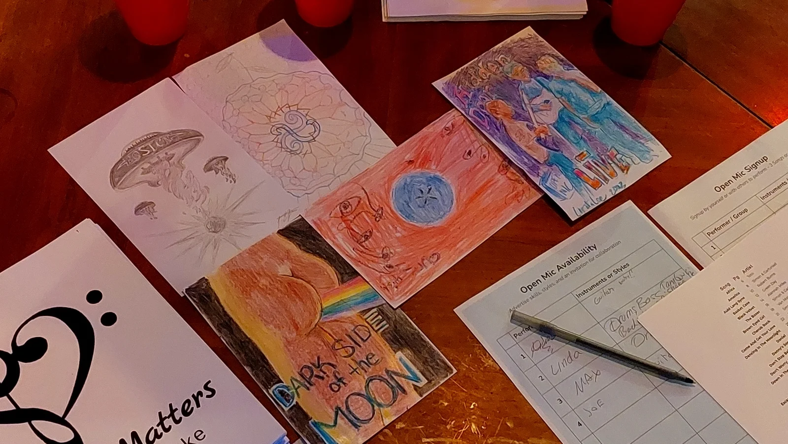 Tabletop display of participant record cover artworks with open mic signup sheets and candles